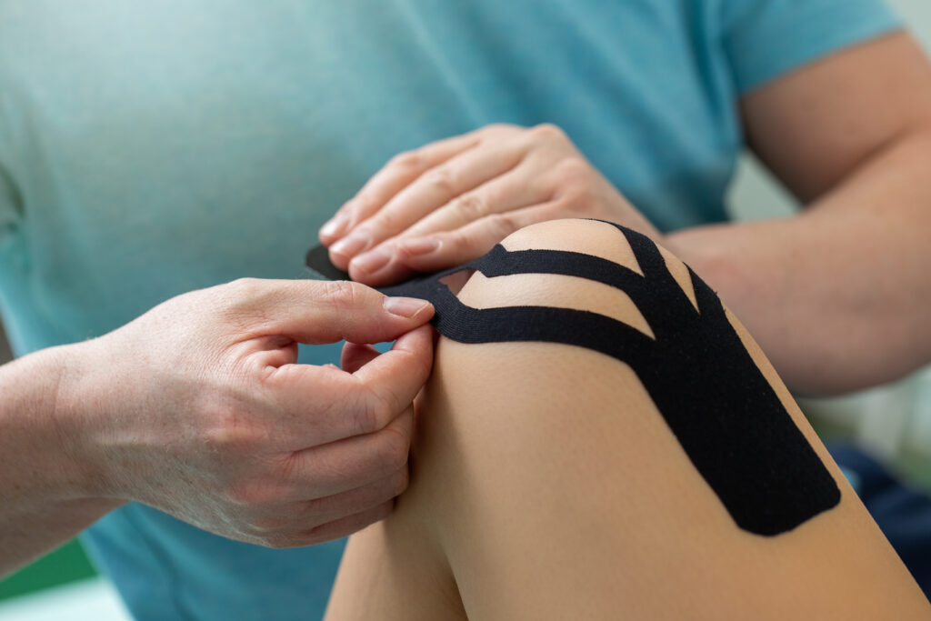 Physiotherapist applying kinesiology tape to patient knee. Therapist treating female Caucasian athlete with a functional bandage. Post traumatic rehabilitation and sport physical therapy.